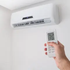 Dłoń trzymająca pilot do klimatyzacji na tle białej ściany z jednostką klimatyzacyjną.