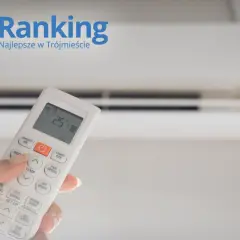 Dłoń trzymająca pilot do klimatyzacji przed białym urządzeniem.