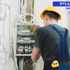Elektryk w żółtym kasku pracuje przy rozdzielnicy, dokonując doboru zabezpieczeń nadprądowych. WN6.