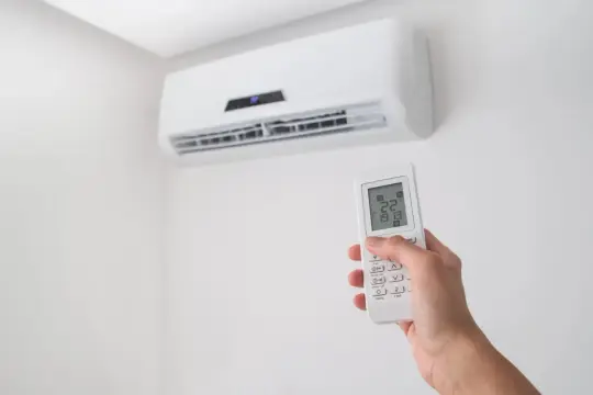 Dłoń trzymająca pilot do klimatyzacji na tle białej ściany z jednostką klimatyzacyjną.