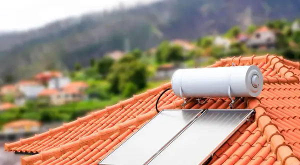 Dwa panele kolektorów solarnych i bojler na dachu z pomarańczowej dachówki, w tle zielone wzgórza i zabudowania.