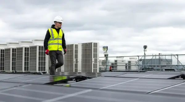Pracownik w kamizelce odblaskowej przechodzi obok rzędu klimatyzatorów na dachu, gdzie instalowane są panele słoneczne. sofar solar polska.