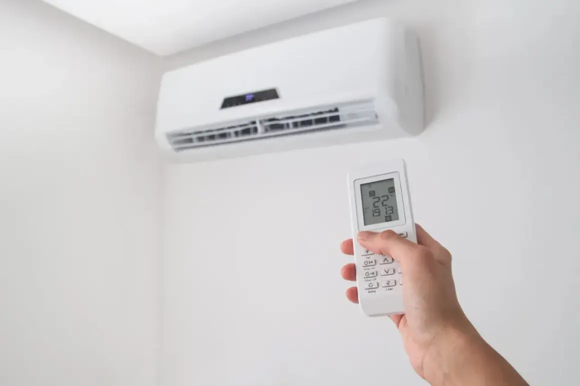 Dłoń trzymająca pilot do klimatyzacji na tle białej ściany z jednostką klimatyzacyjną.
