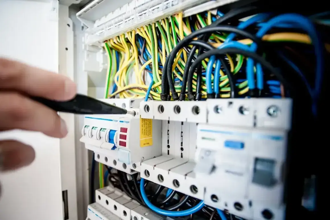 Ręka majstruje przy bezpiecznikach, gdzie kolorowe przewody symbolizują przepływ prądu elektrycznego.