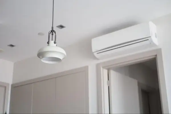 Biała klimatyzacja ścienna nad drzwiami, obok lampy wiszącej.